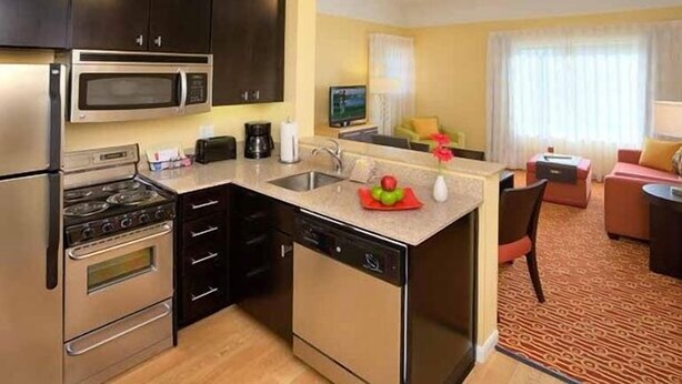 A compact kitchen with a microwave, stove, and dishwasher, a small dining area, and a living room with a sofa and a TV. - Towneplace Suites by Marriott Aiken Whiskey Road (Aiken)