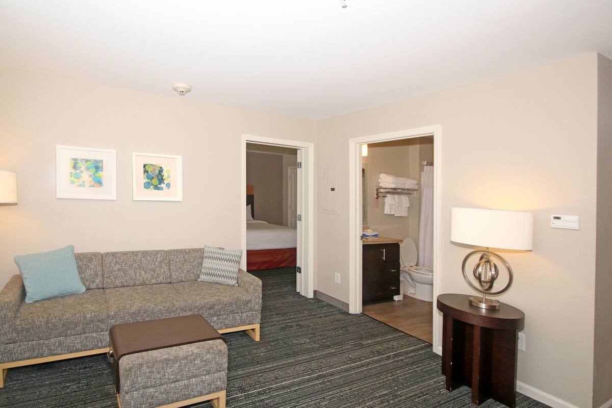 Ein Hotelzimmer mit grauem Sofa, einem kleinen Hocker, einer Lampe und zwei gerahmten Kunstwerken an der Wand.