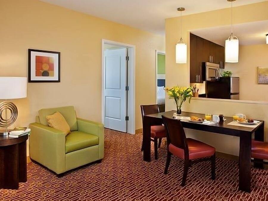 Habitación de hotel con comedor, un sillón verde, una mesa dispuesta para el desayuno y una cocina visible a través de una puerta abierta.
