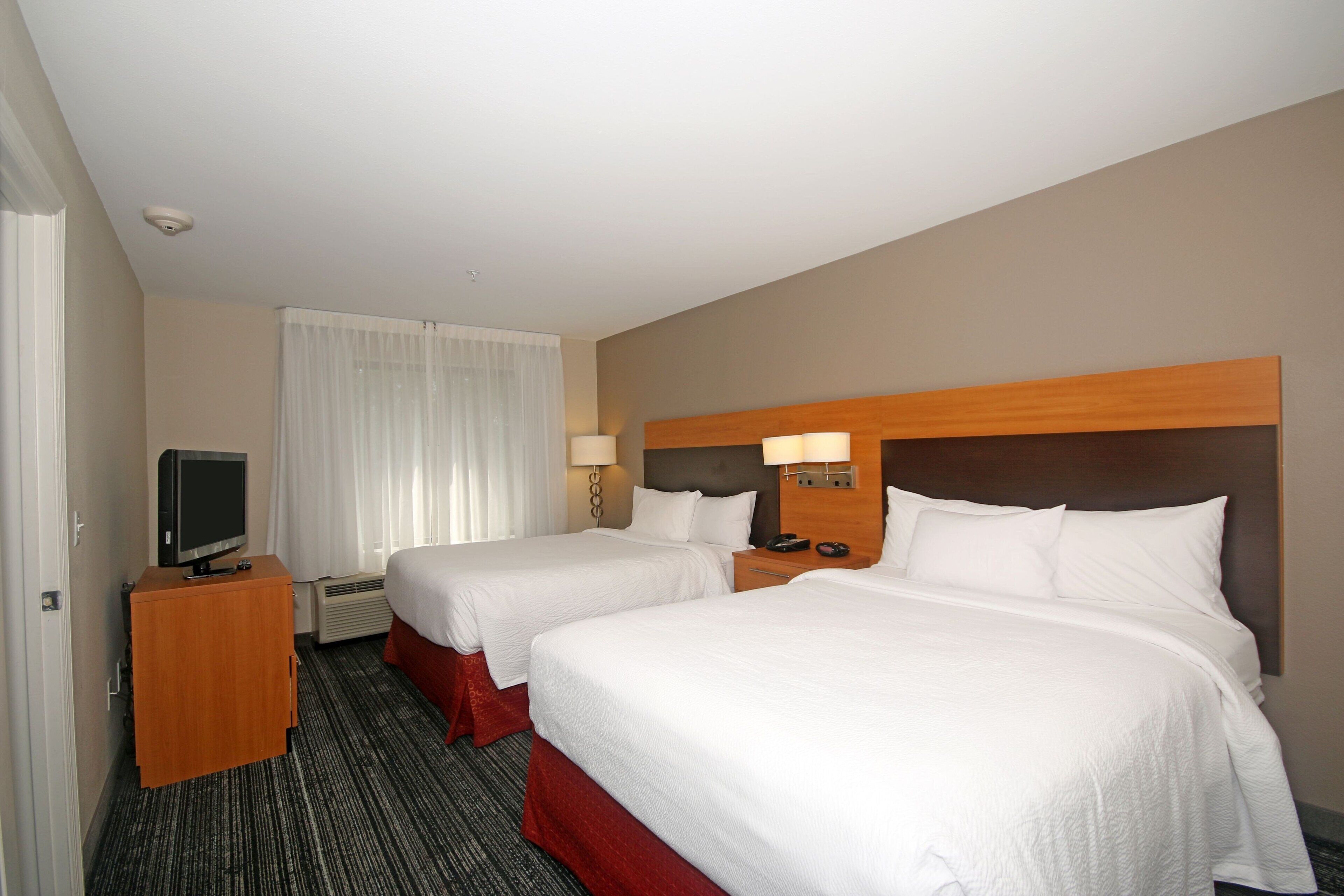 A hotel room with two beds, a television on a wooden cabinet, and a window with white curtains.