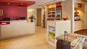 A hotel reception area with a reception desk, a 'Hello' sign, a seating area with a table and chairs, a bookshelf with magazines, and a small kitchenette. - Towneplace Suites by Marriott Aiken Whiskey Road (Aiken)