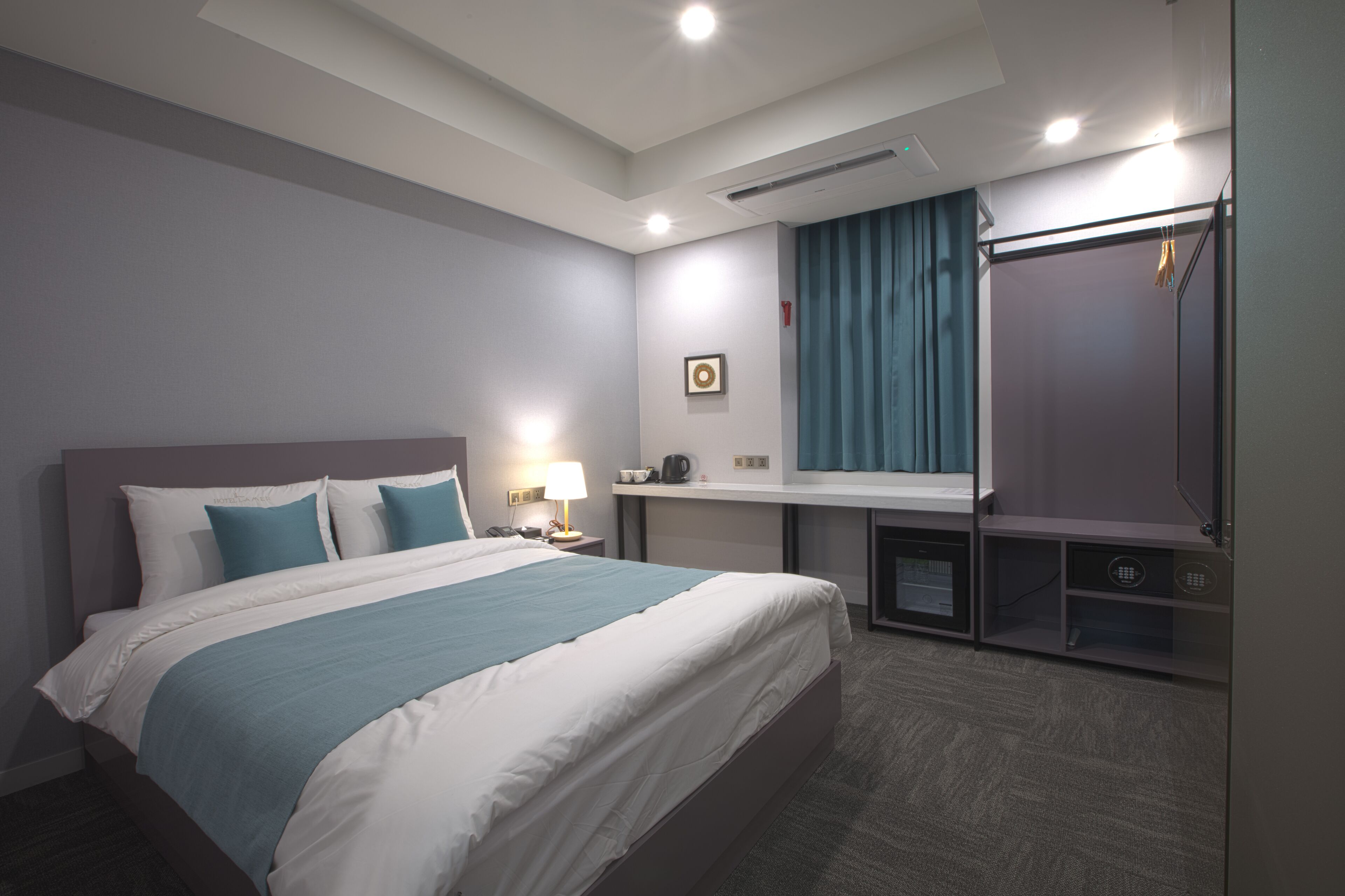 A hotel room with a large bed, a desk with a lamp, a mirror, a wardrobe, and a small kitchenette.
