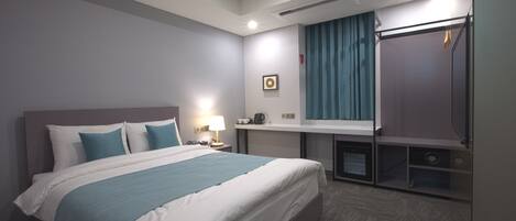 A hotel room with a large bed, a desk with a lamp, a mirror, a wardrobe, and a small kitchenette.