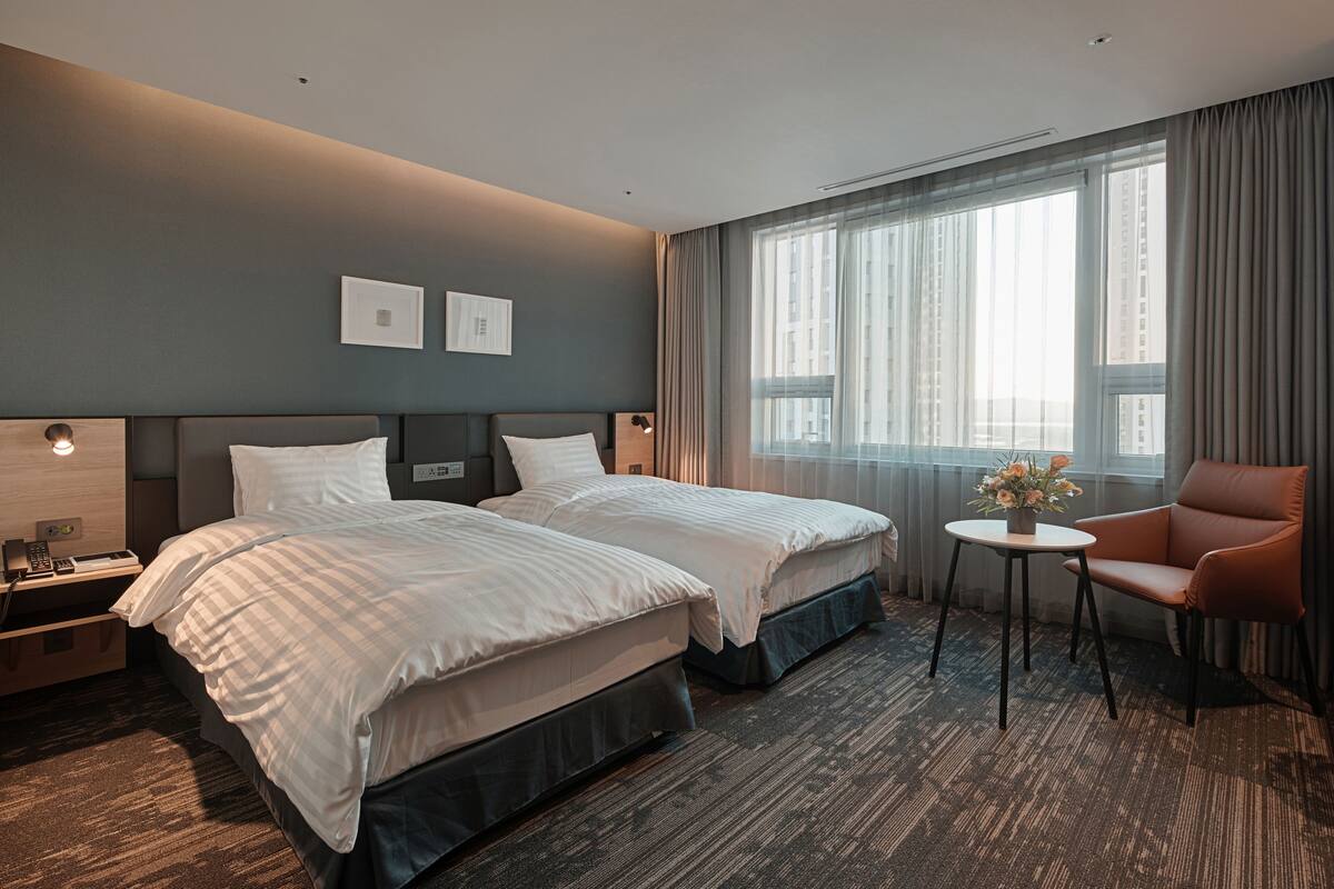 A hotel room with two beds, a small table with a vase of flowers, a chair, and a large window with curtains.