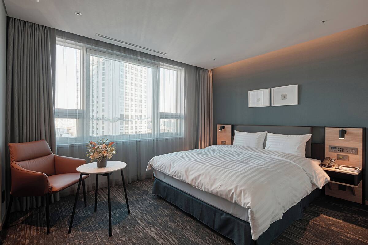 A hotel room with a bed, a chair, a small table with a vase of flowers, a nightstand with a phone, and a view of a cityscape through large windows.