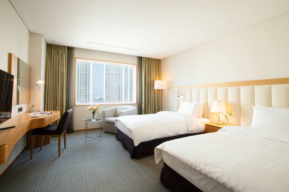 A hotel room with two beds, a desk, a chair, a small table with a vase, and a window with curtains.