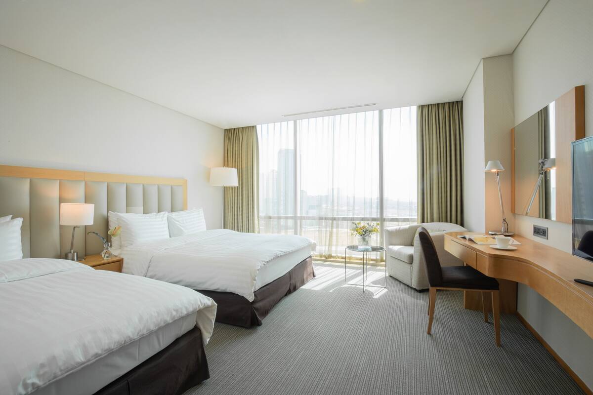 A hotel room with two beds, a desk, a chair, a TV, and a large window with curtains.