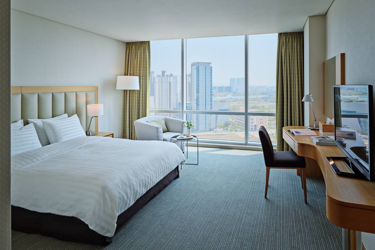 A hotel room with a large bed, a desk with a computer, a chair, and a view of the cityscape through large windows.