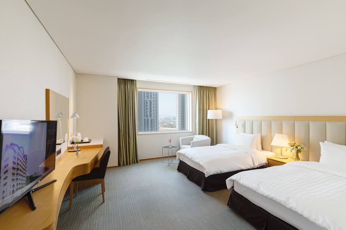 A hotel room with two beds, a desk with a TV, a small table with a lamp, and a window with curtains.