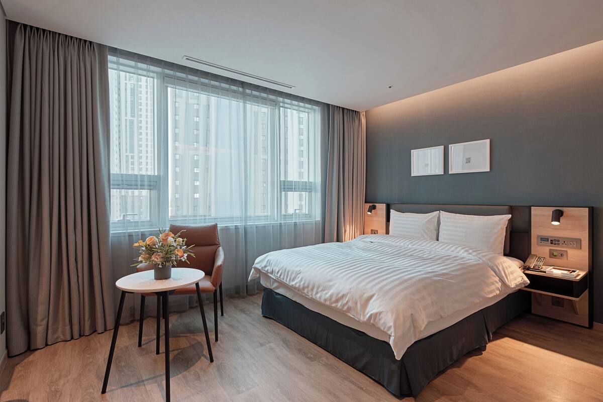A modern hotel room with a large bed, a small table with a vase of flowers, a chair, and a nightstand with a phone and lamp.