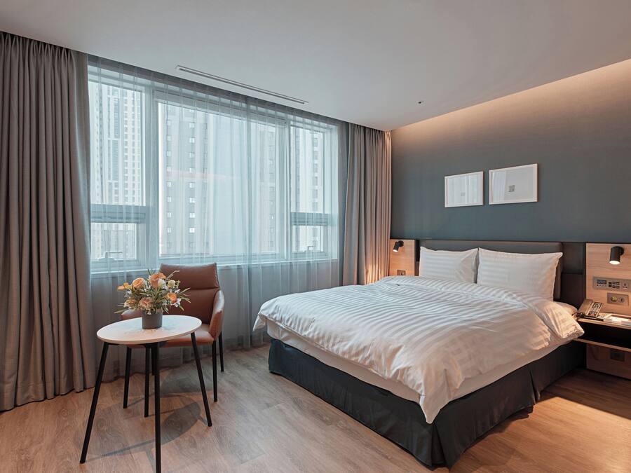 Une chambre d’hôtel moderne avec un grand lit, une petite table ornée d’un vase de fleurs, une chaise et une table de chevet avec un téléphone et une lampe.