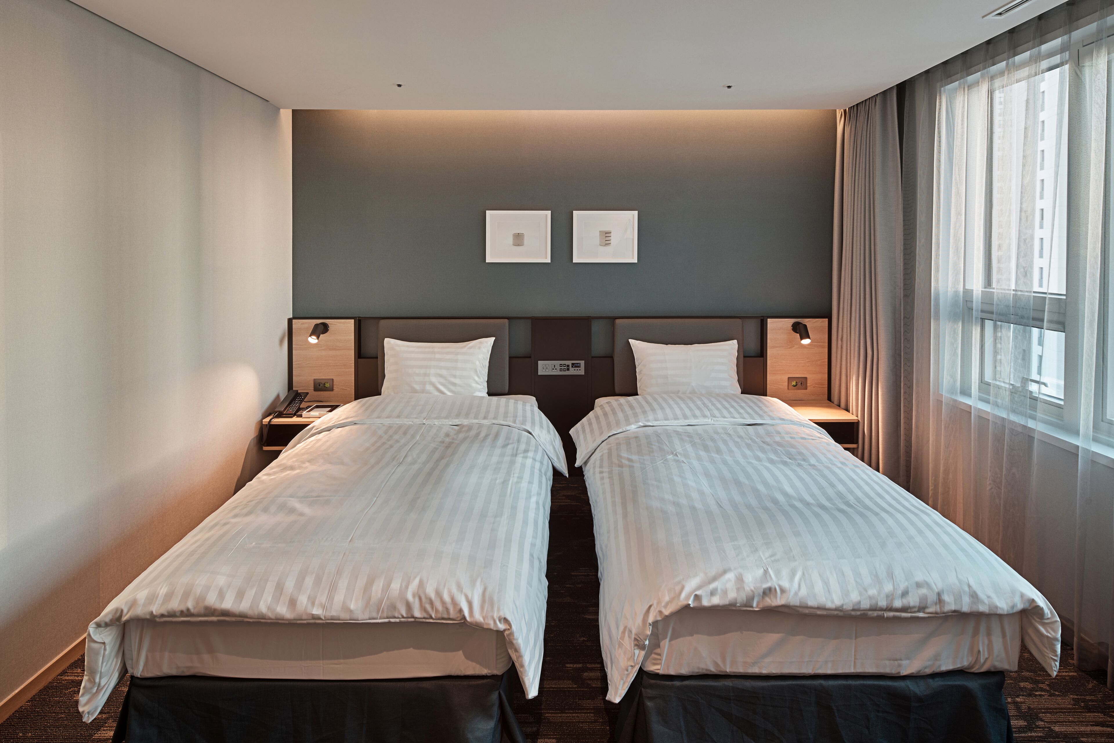 A hotel room with two single beds, a nightstand with a lamp, a wall-mounted control panel, and a window with curtains.