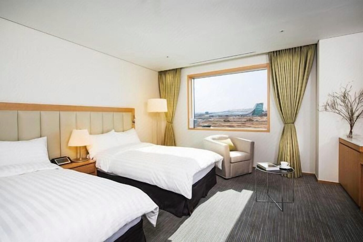 A hotel room with two beds, a desk, a chair, a window with a view, and a small table with a lamp.