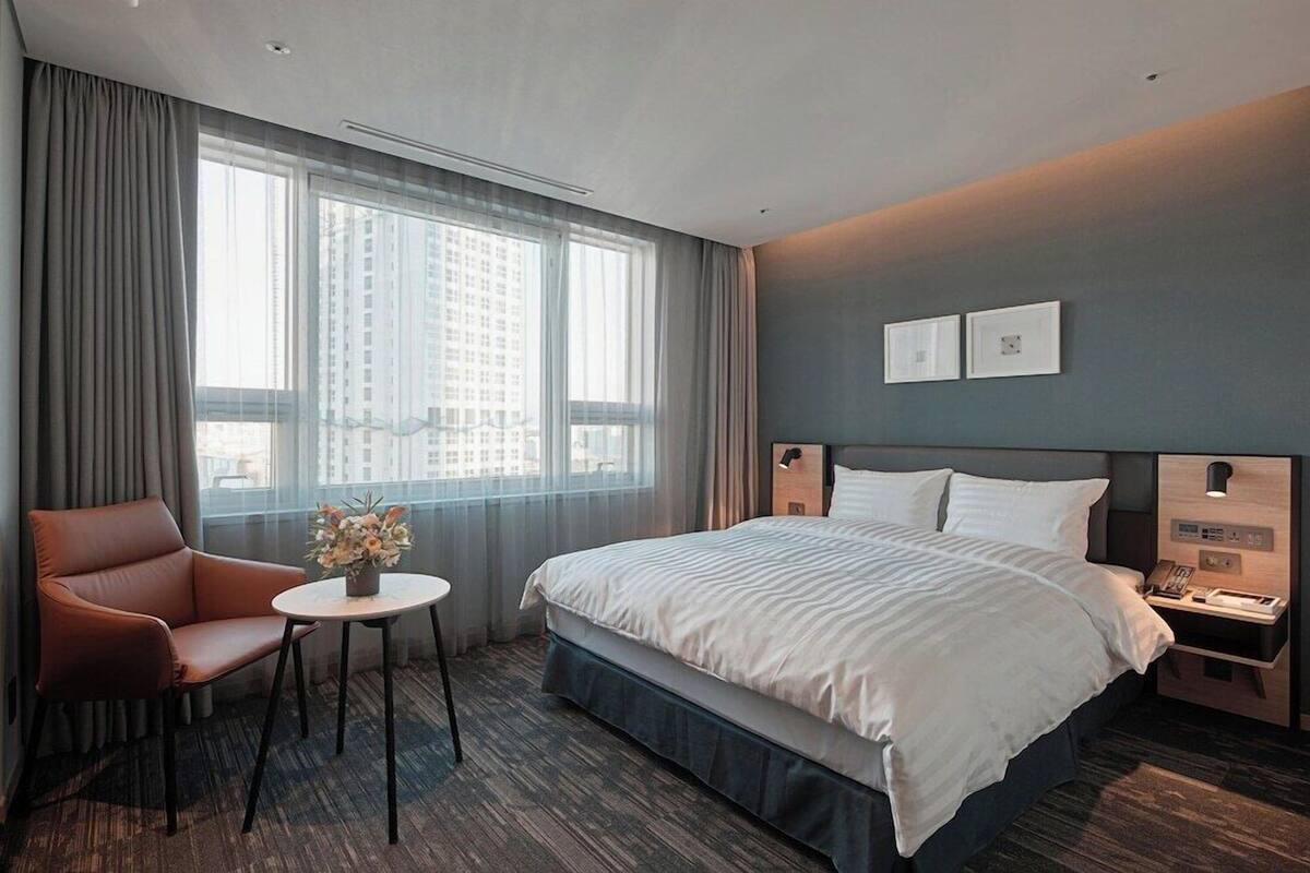 A hotel room with a bed, a chair, a small table with flowers, a nightstand, and a view of the city.