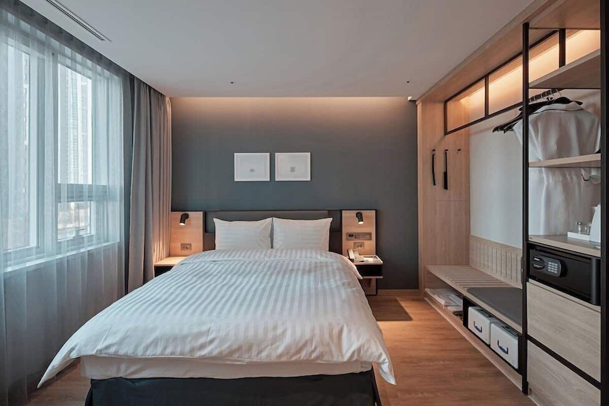 A modern hotel room with a large bed, bedside tables, a wardrobe, and a view of the city.