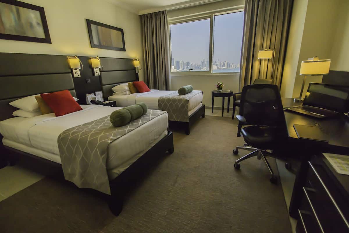 A hotel room with two beds, a desk, a chair, and a view of the city.