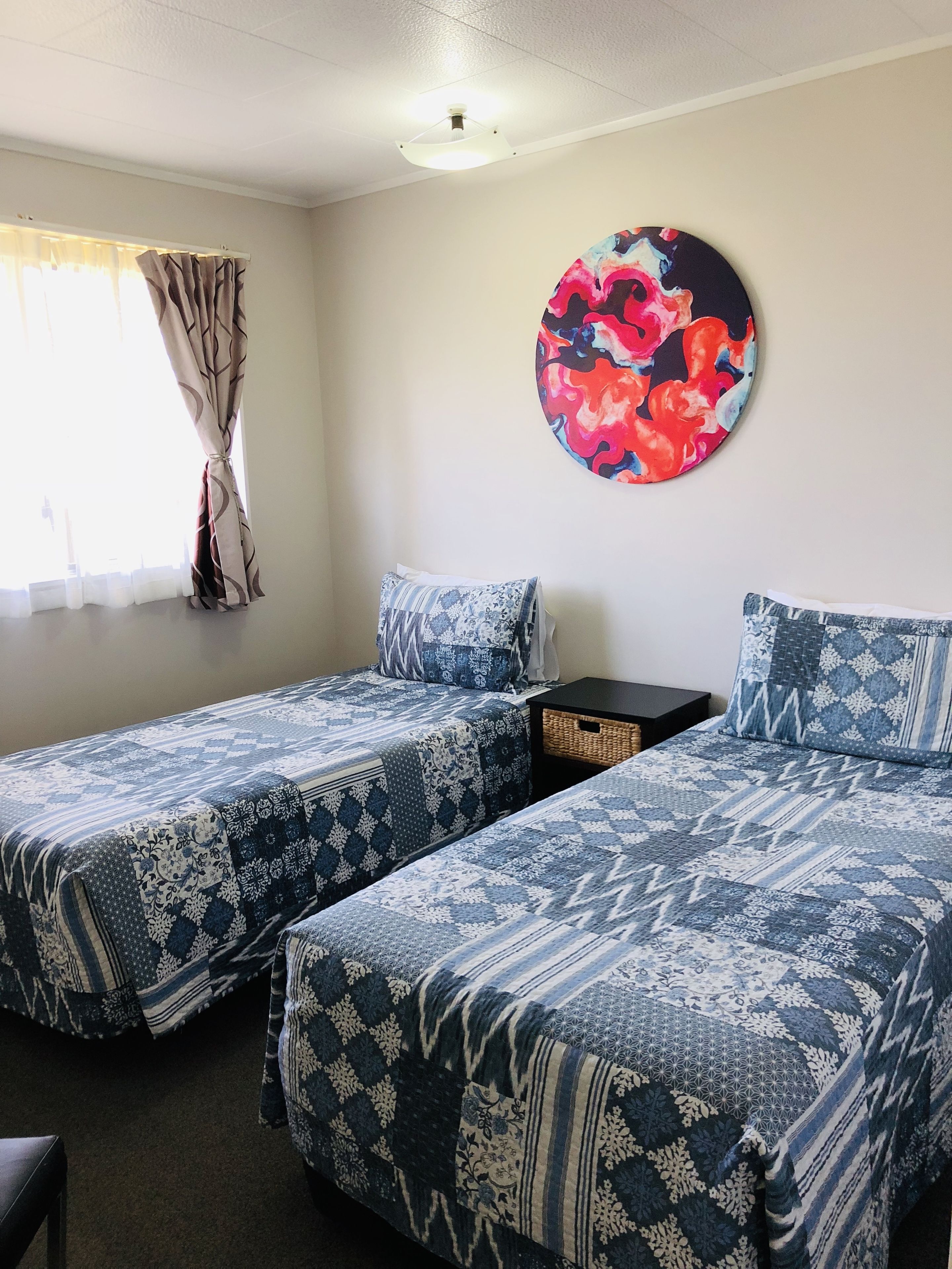Une chambre avec deux lits, une petite table et un tableau abstrait aux couleurs vives accroché au mur.