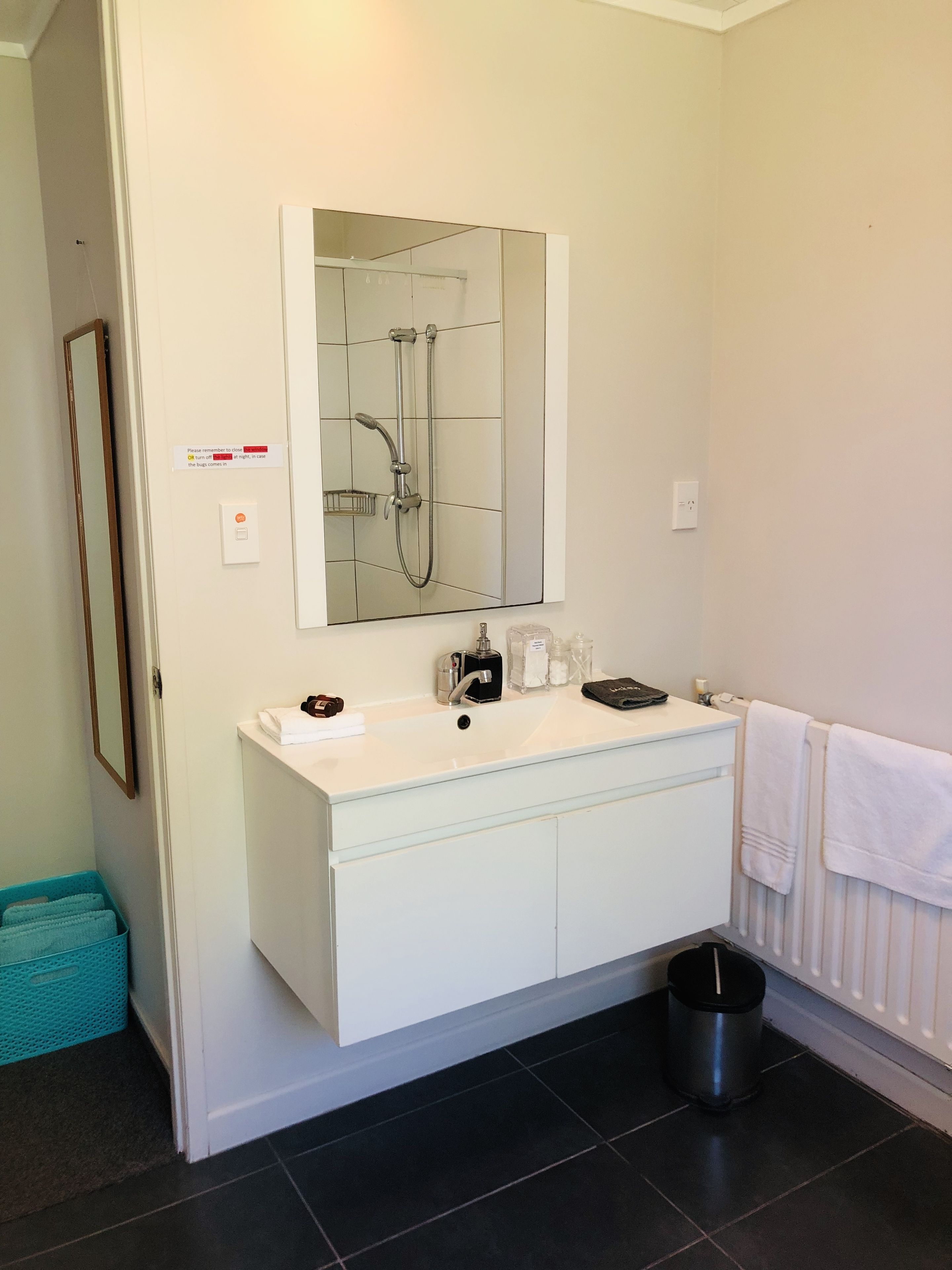 Ein modernes Badezimmer mit einem weißen Waschtisch, einem großen Spiegel, einer Dusche und einem schwarzen Mülleimer.