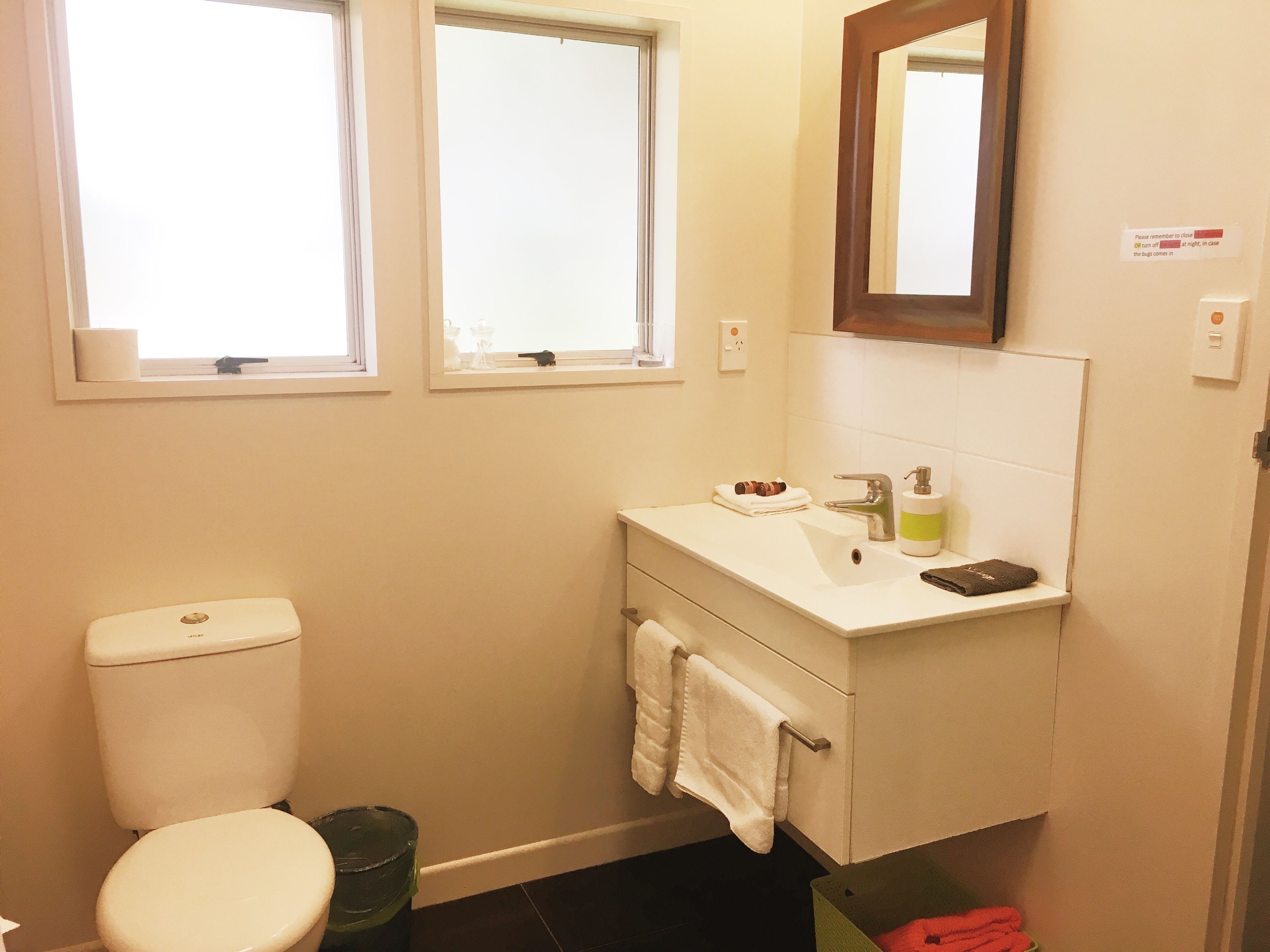 Ein kleines, sauberes Badezimmer mit Toilette, Waschbecken und Spiegel. Zwei Fenster sorgen für Tageslicht.