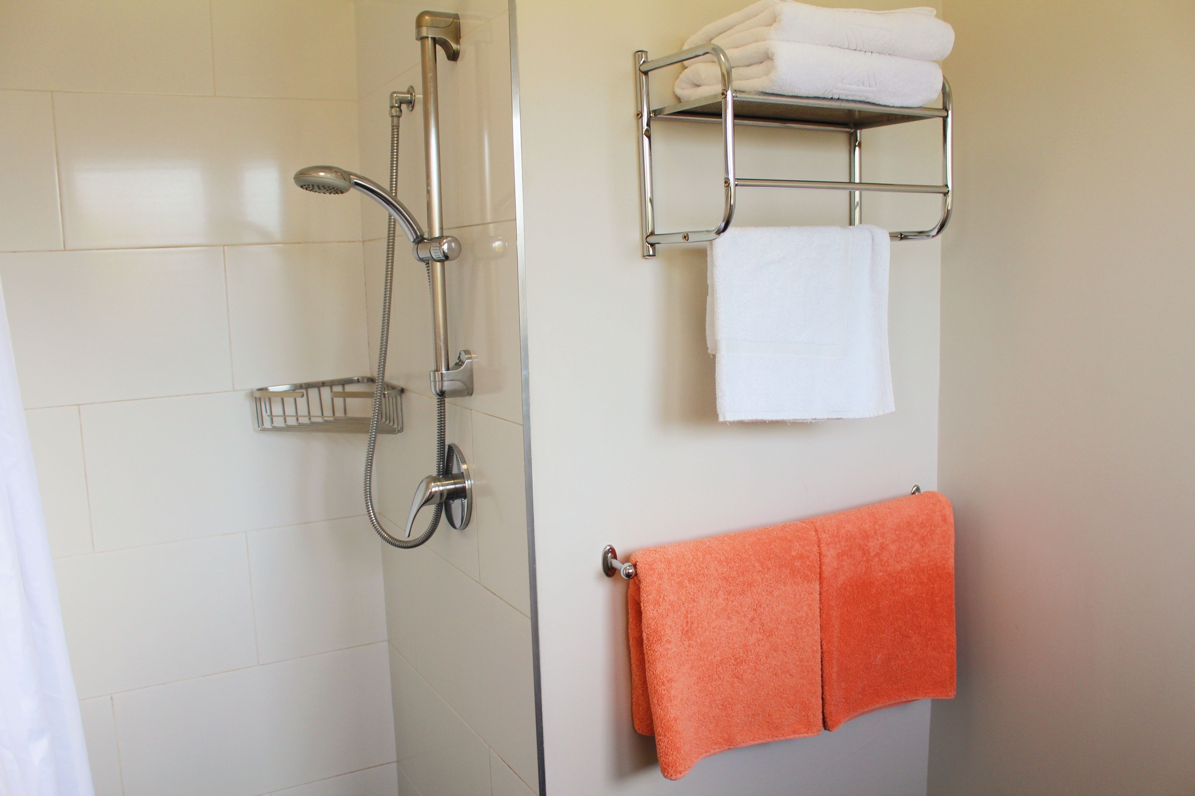 Un baño con ducha, un perchero con toallas blancas y anaranjadas, y una estantería metálica con toallas blancas.