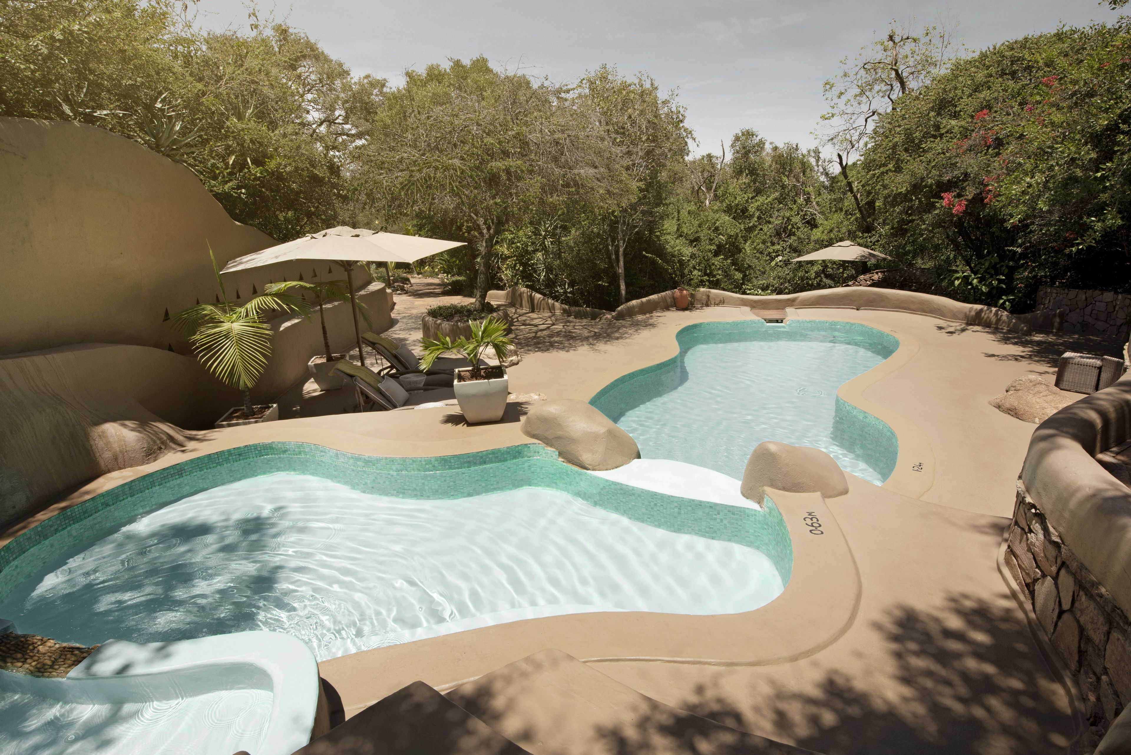 Una piscina con diseño ondulado, rodeada de sillas de descanso y sombrillas, ubicada en un área ajardinada con árboles y plantas.