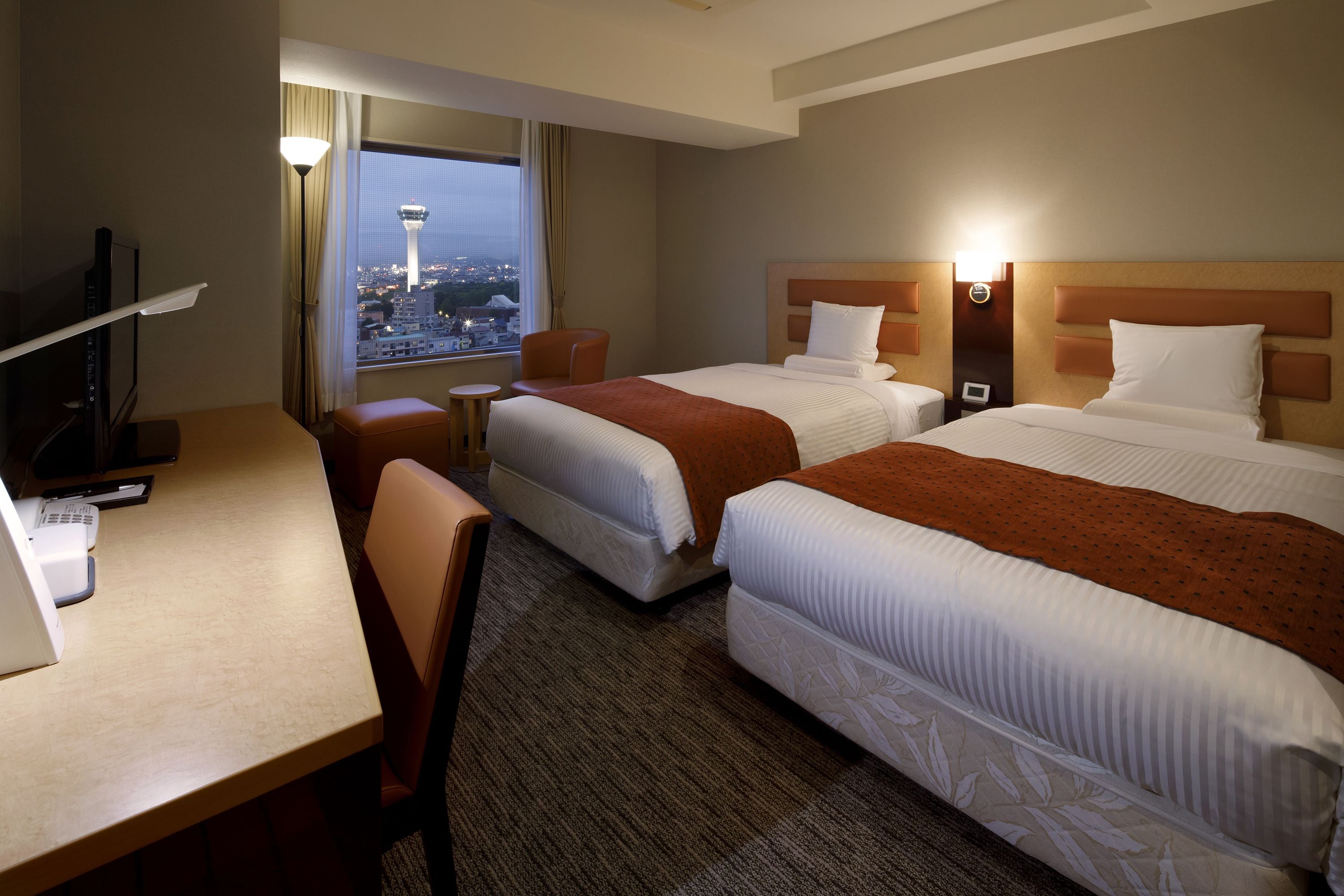 A hotel room with two beds, a desk, a chair, and a view of a cityscape.