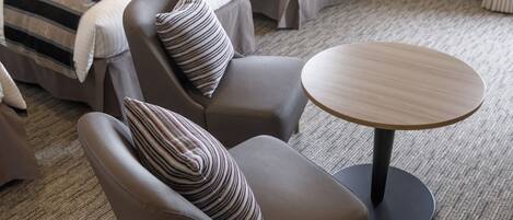 A hotel room with two grey armchairs, a round wooden table, and a bed with striped bedding.