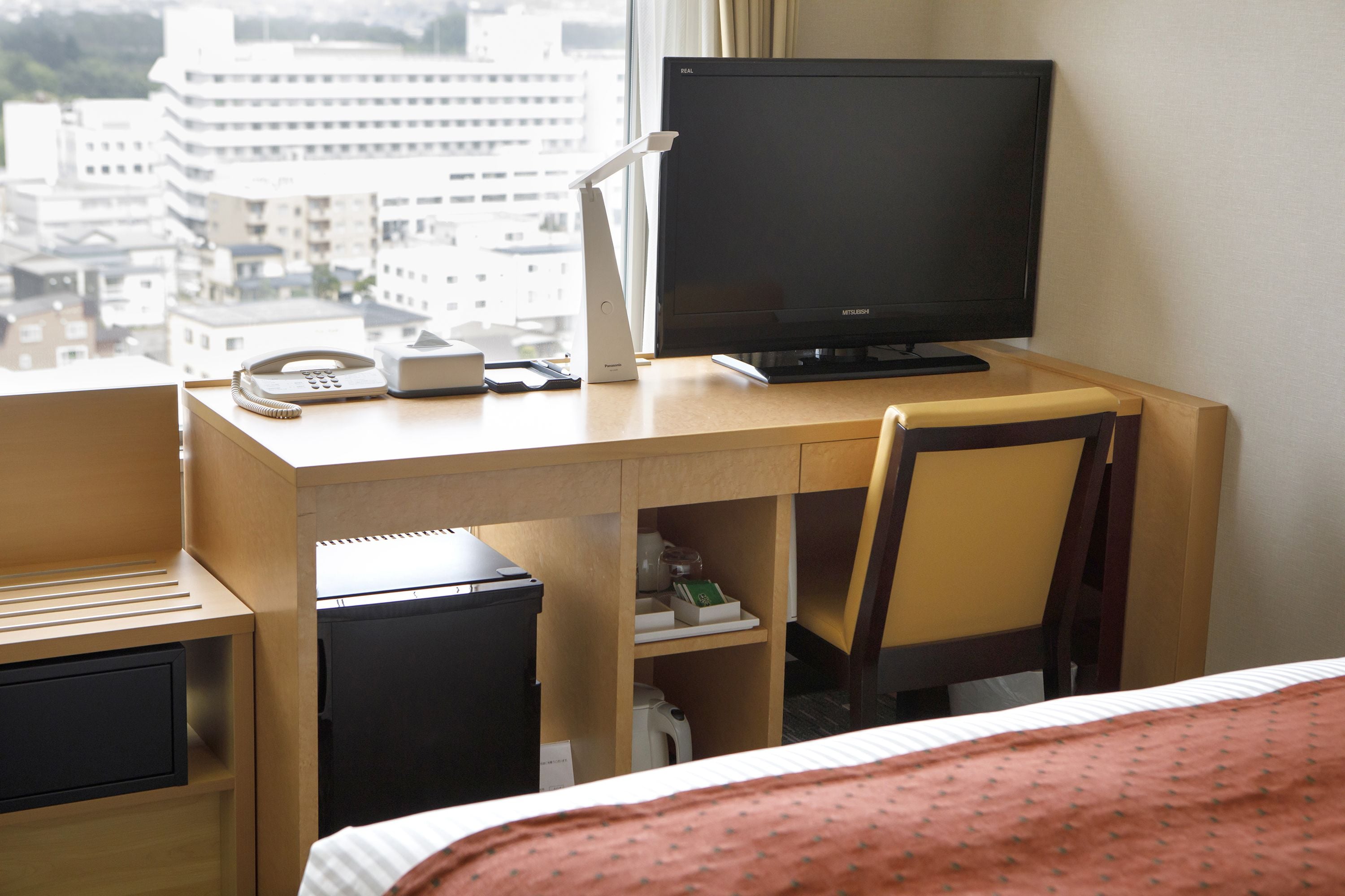 Une chambre d’hôtel avec un bureau en bois, un ordinateur, un téléphone et un lit recouvert d’une couverture rouge.
