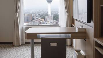A hotel room with a view of a cityscape and a tower, a desk, a chair, and a TV mounted on the wall.
