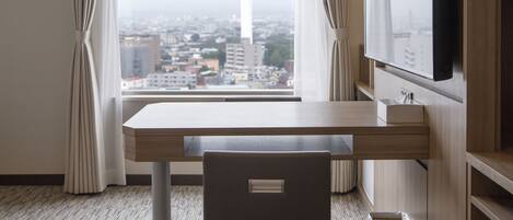A hotel room with a view of a cityscape and a tower, a desk, a chair, and a TV mounted on the wall.