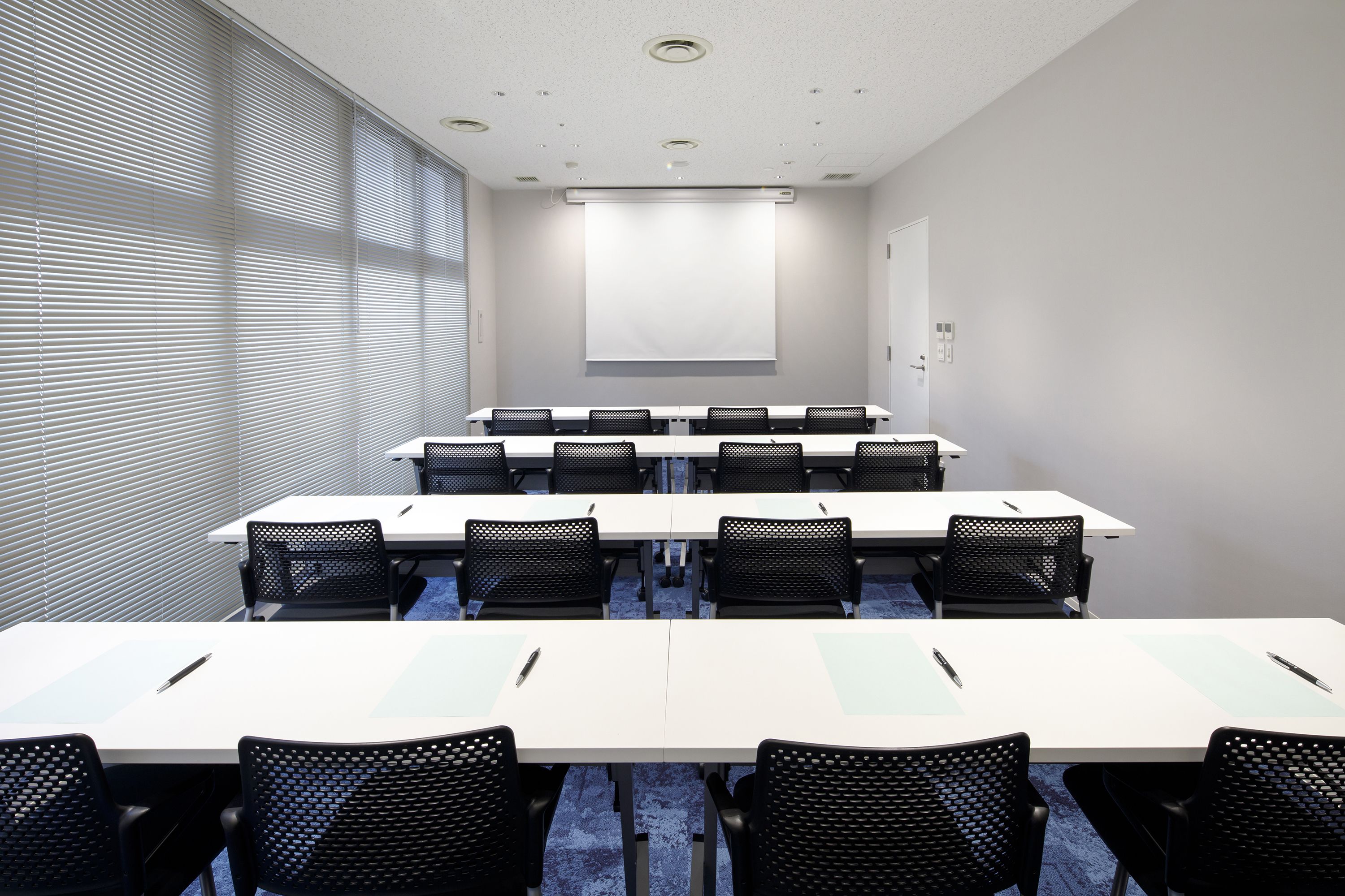 A conference room with a projector screen, white tables, and black mesh chairs.