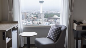 Ein Hotelzimmer mit Blick auf die Stadtlandschaft, einem kleinen runden Tisch, einem gepolsterten Sessel und einem Schreibtisch mit Telefon.