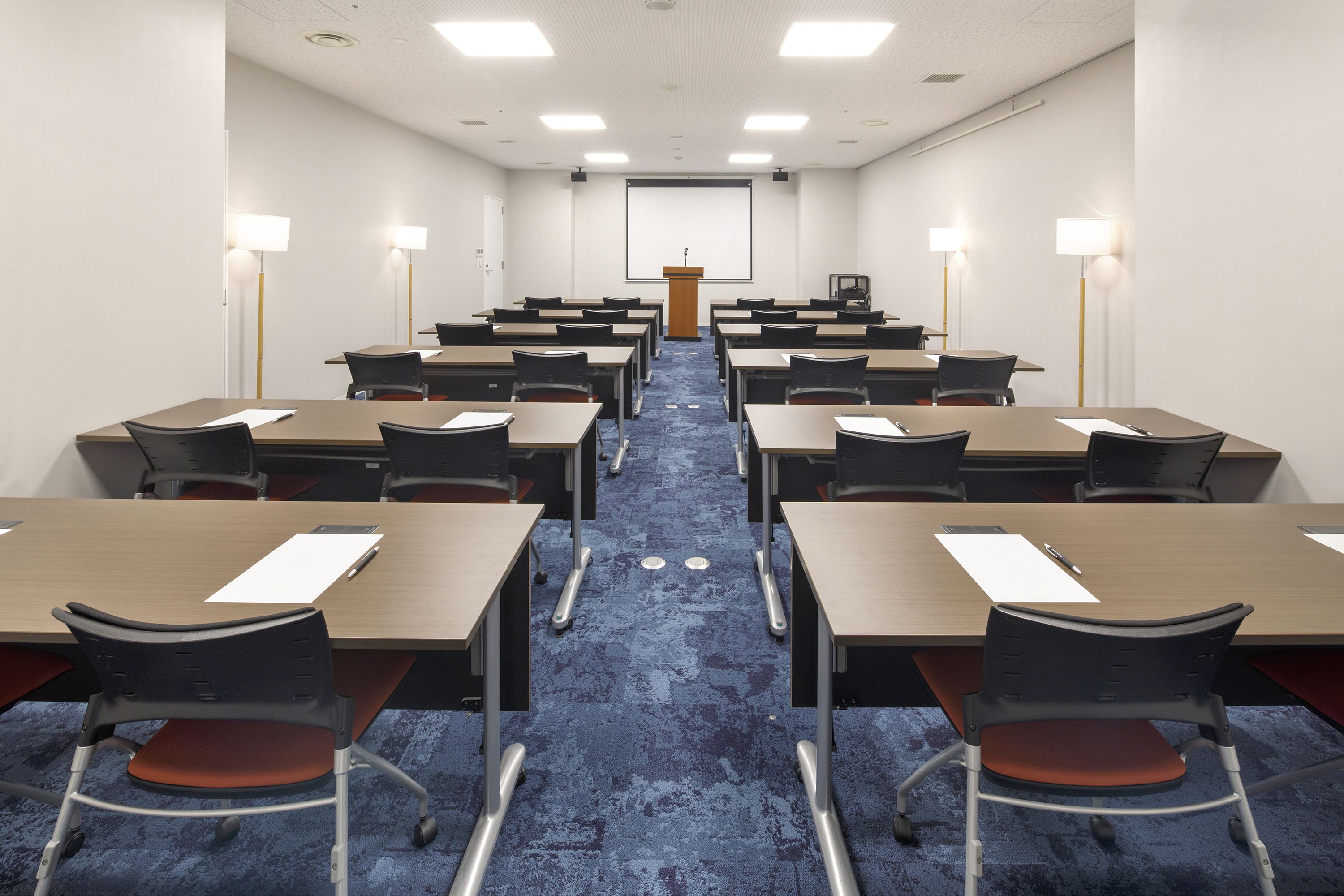 A conference room with rows of tables and chairs, a podium, and wall-mounted lamps.