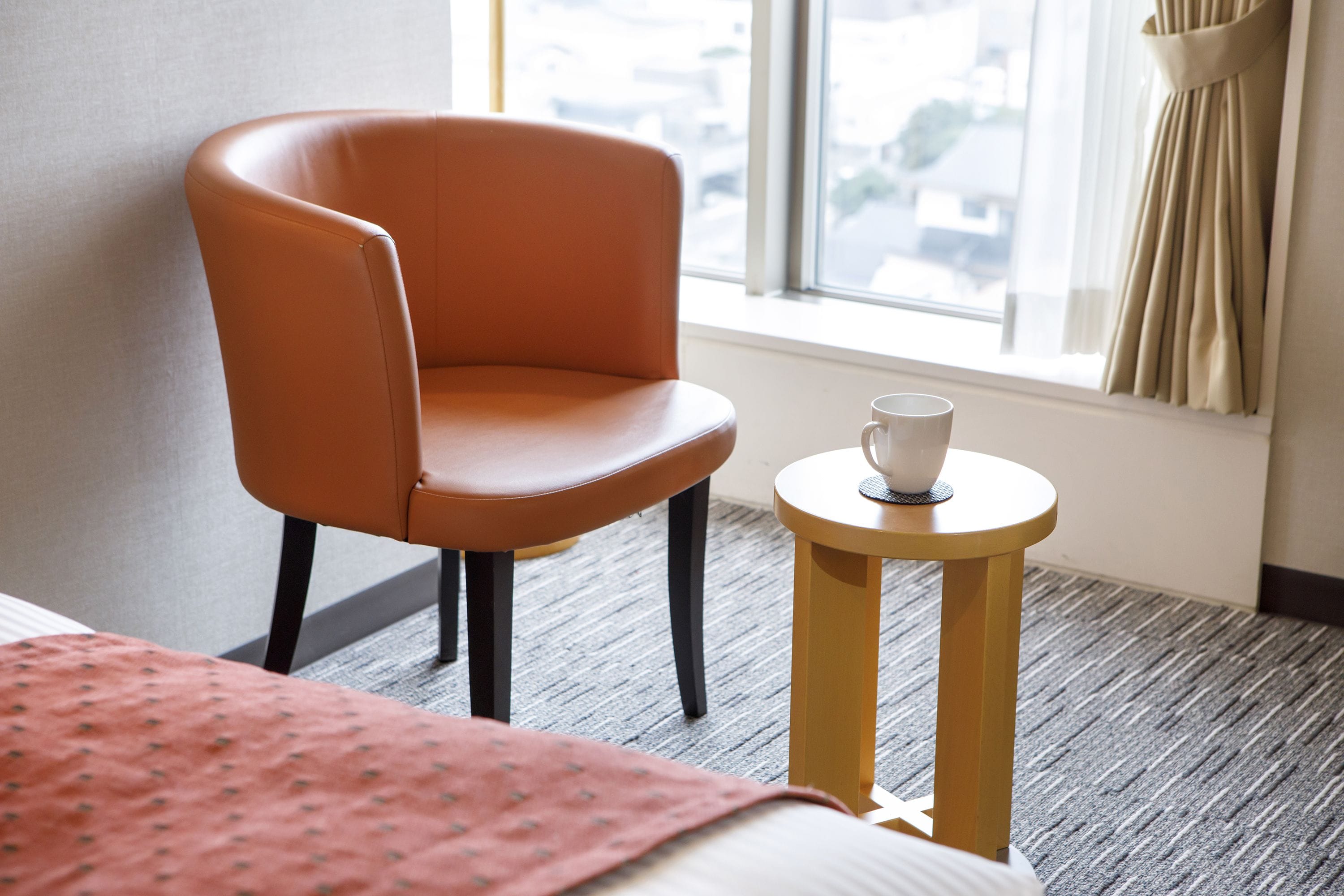 A hotel room with a single orange chair, a small round table with a cup, and a bed with a red blanket.
