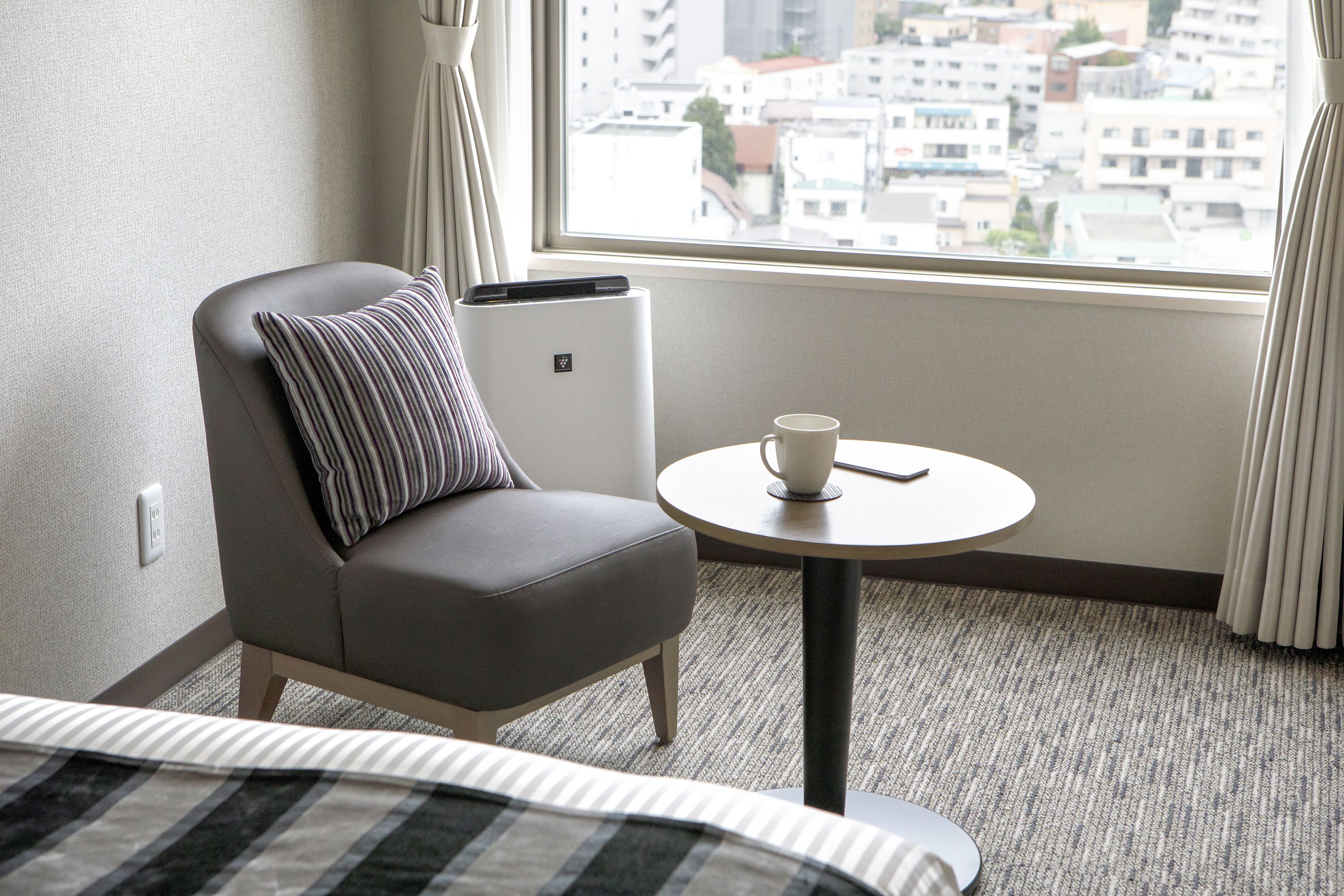 Un cuarto de hotel con una cama, un sillón gris con un cojín a rayas, una mesita redonda con una taza, y vistas de edificios a través de una ventana.
