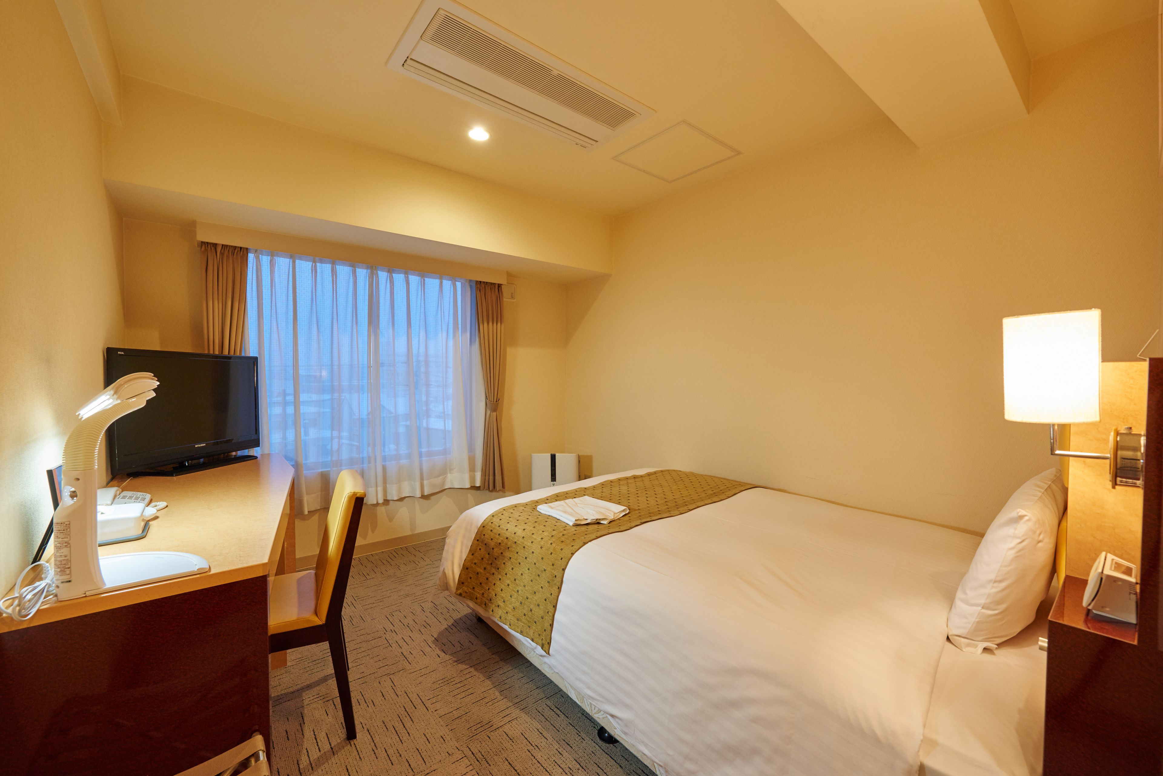 A hotel room with a bed, a desk with a computer, a television, and a window with curtains.