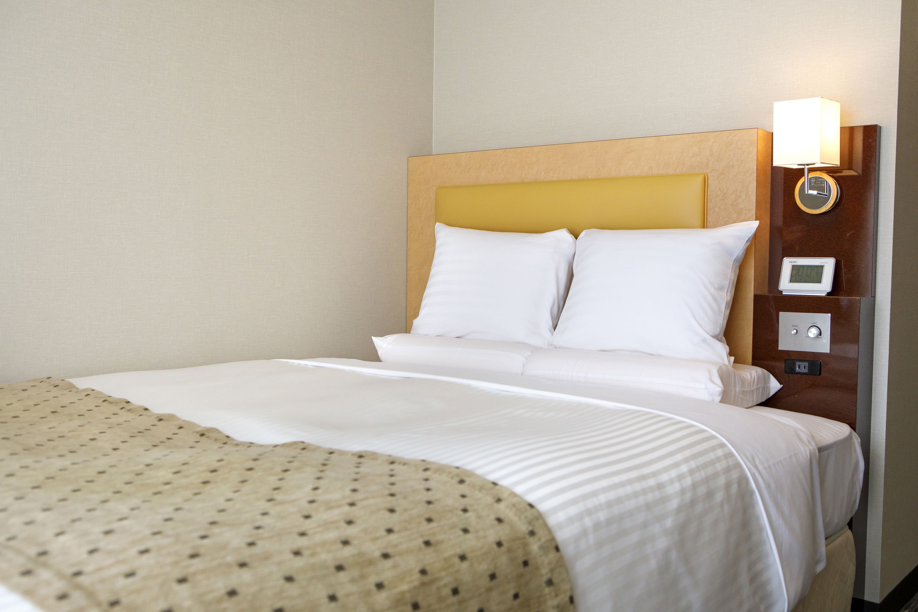 A hotel room with a large bed, a headboard, a bedside table with a lamp, and a control panel.