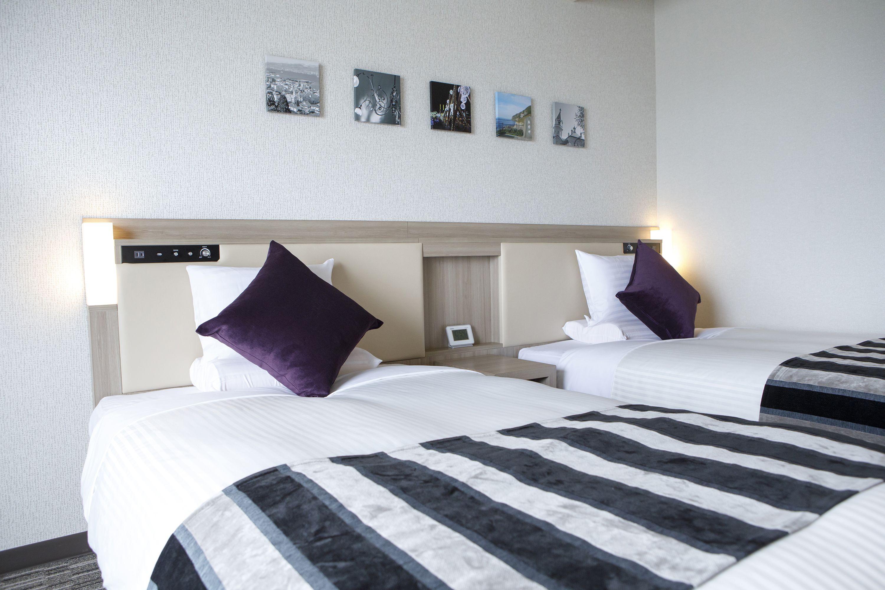 A hotel room with two beds, each with a headboard, white bedding, and a striped blanket. There are wall-mounted picture frames and a small bedside table with a lamp.