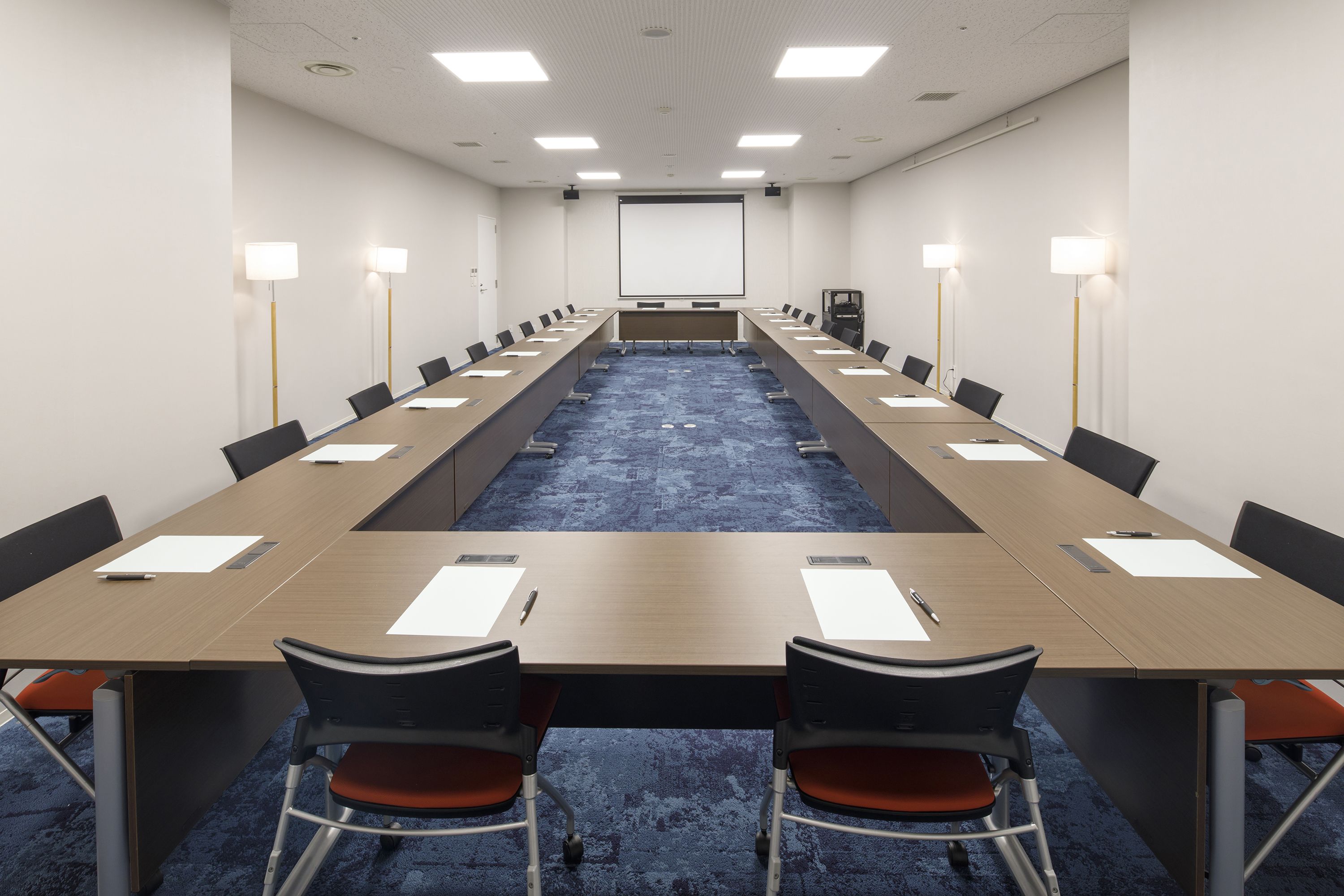 A conference room with a long table, chairs, a projector screen, and a carpeted floor.