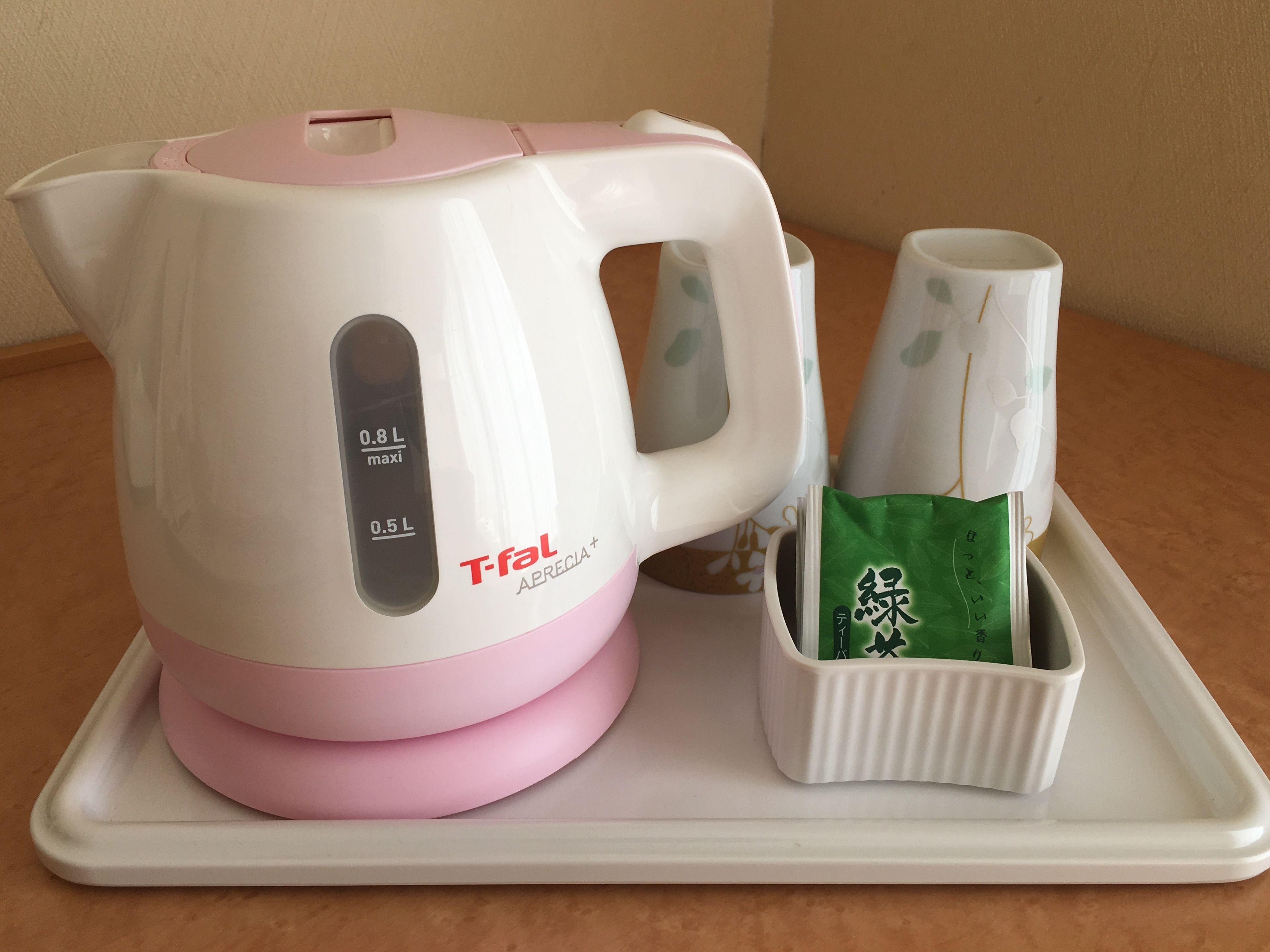 Une bouilloire électrique blanche et rose dotée d’un écran numérique, un sachet de thé vert et deux tasses blanches ornées de motifs de feuilles vertes, le tout disposé sur un plateau.