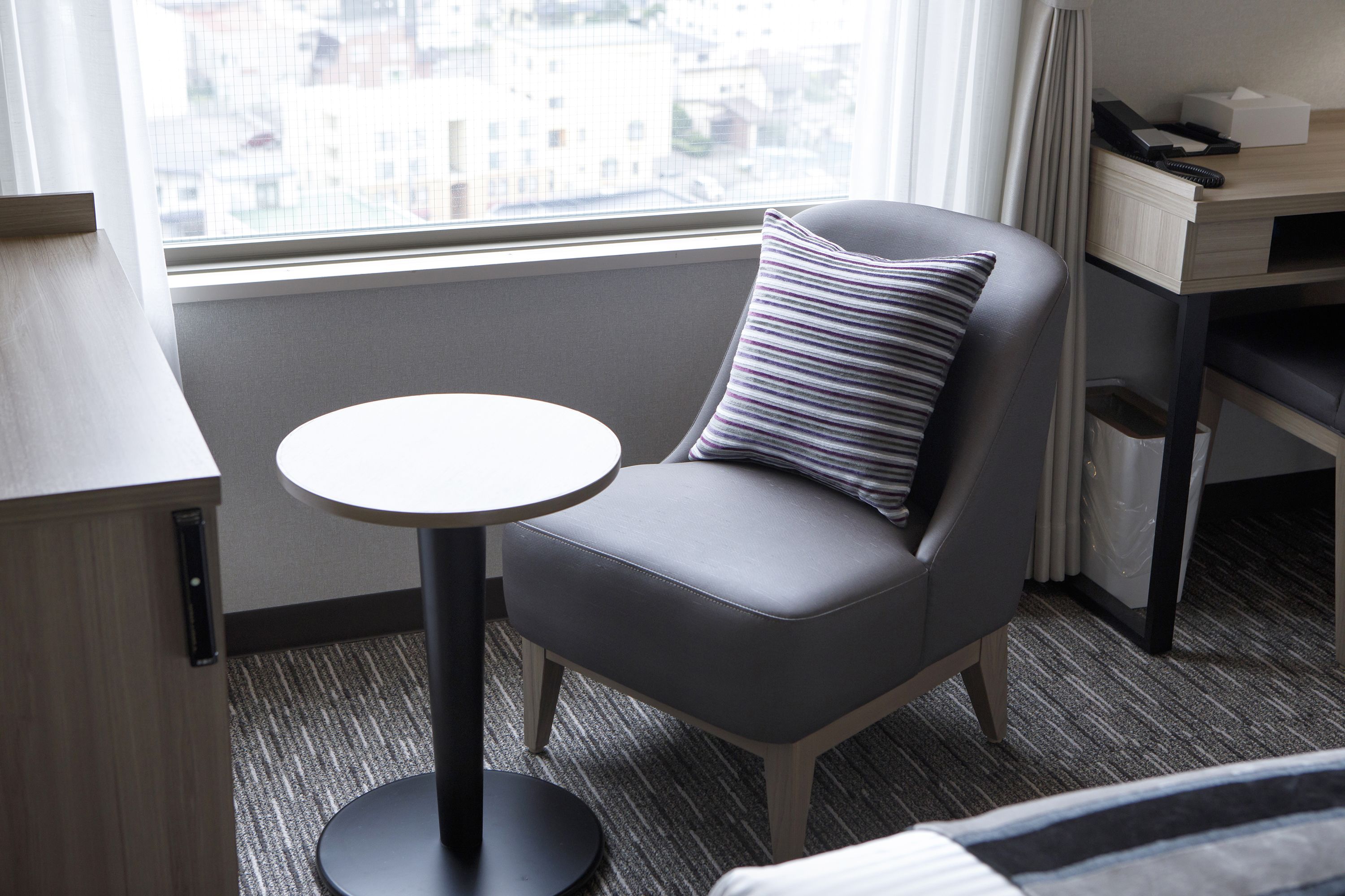 Una habitación de hotel moderna con una butaca tapizada de gris, una mesita redonda y un escritorio con computadora y teléfono.
