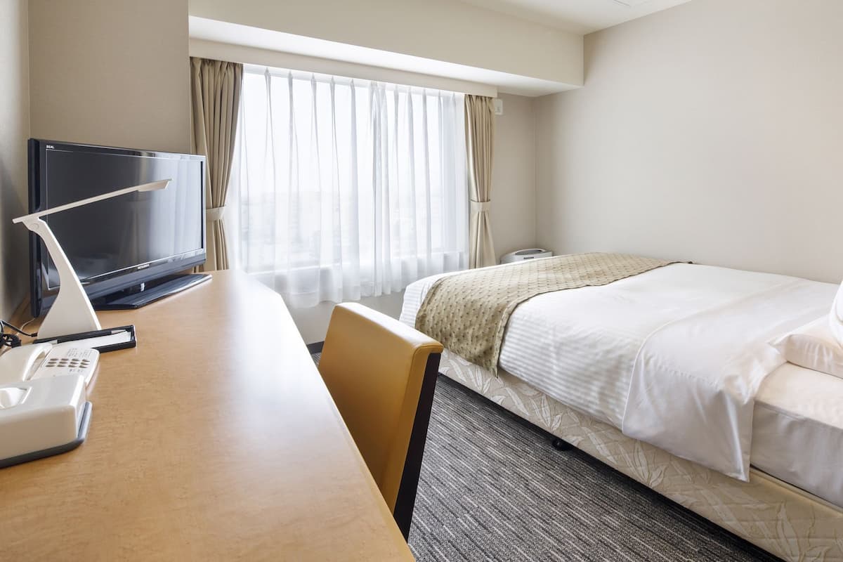 A hotel room with a bed, a desk with a computer and phone, a TV, and a window with curtains.