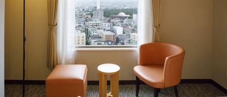 A hotel room with a bed, two chairs, a small round table, and a view of a cityscape through a large window.