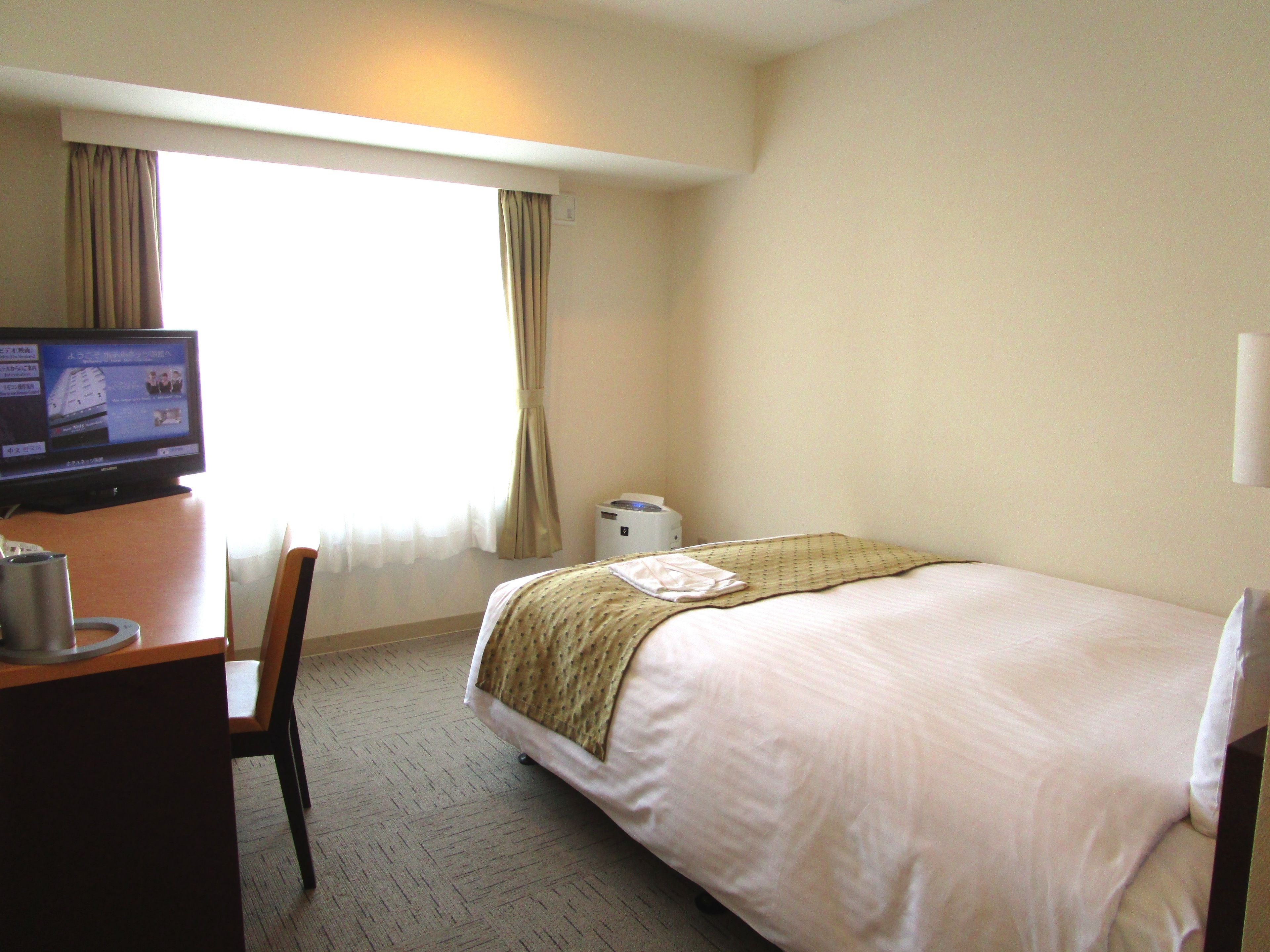 A hotel room with a bed, a desk with a computer, a chair, a window with curtains, and a wall-mounted lamp.