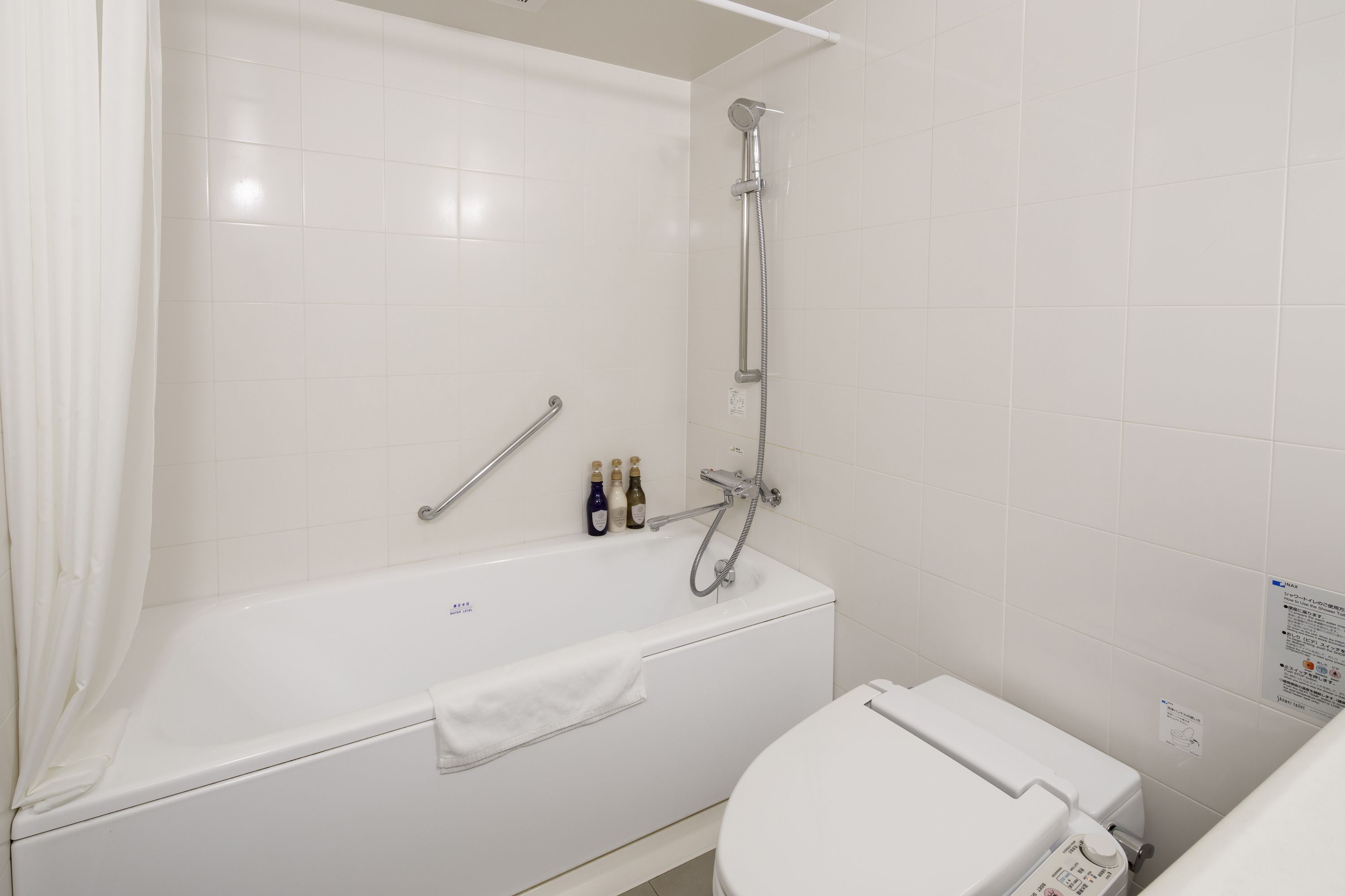 Un baño blanco con bañera, ducha y retrete. Hay botellas de champú y acondicionador en el borde de la bañera.