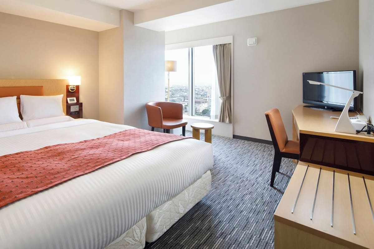 Habitación de hotel con una cama grande, un escritorio con televisor, una silla y ventana con vista a la ciudad.
