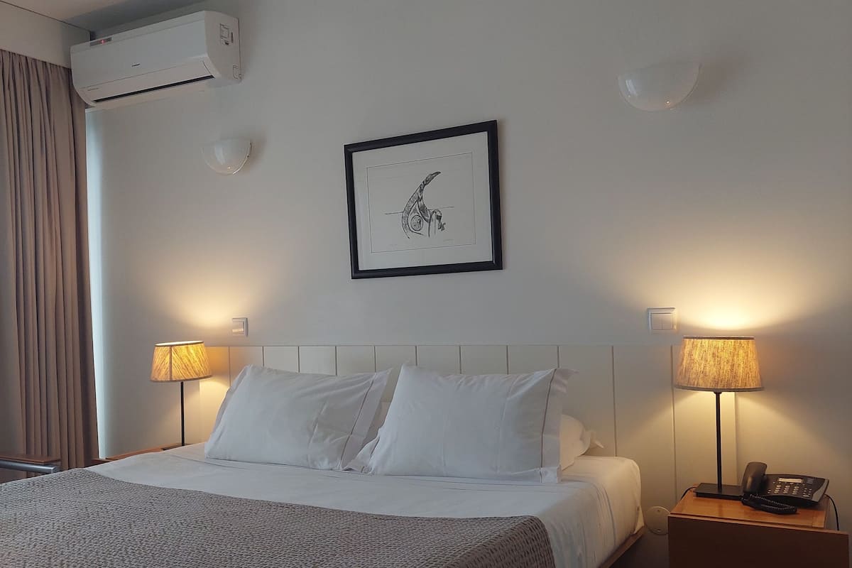 A hotel room with a bed, two bedside lamps, a framed picture on the wall, and an air conditioning unit.