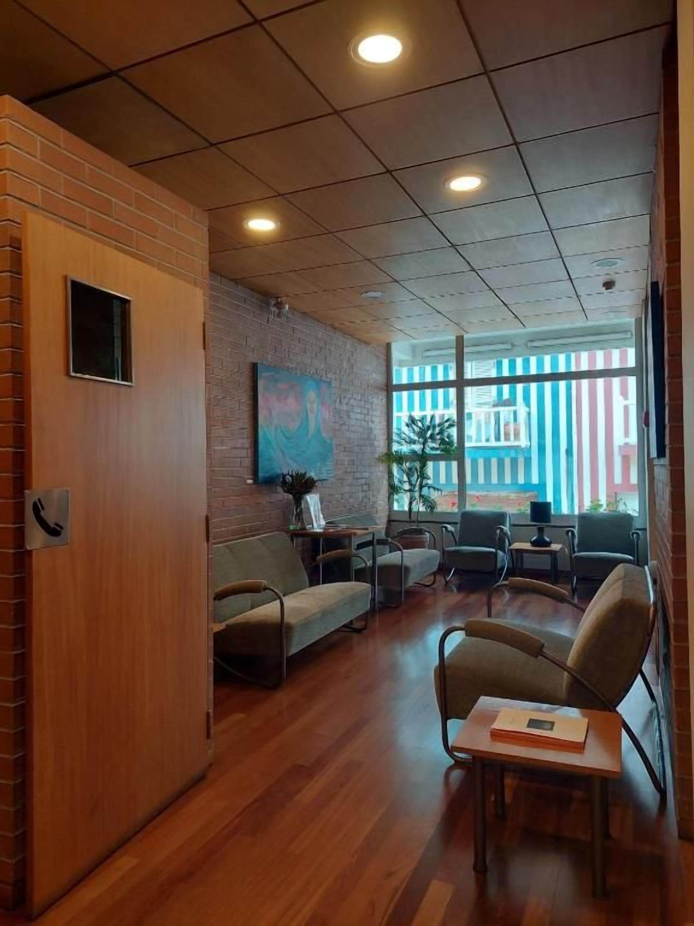 Un área de recepción con un mostrador de madera, una zona de estar con sofás y sillones, un ventanal con vistas a edificios y una pared de ladrillo con un cuadro.