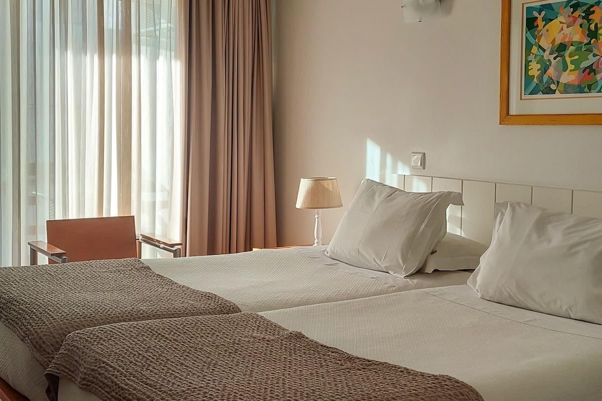 A hotel room with a large bed, a bedside table, a lamp, an air conditioning unit, and a window with curtains.