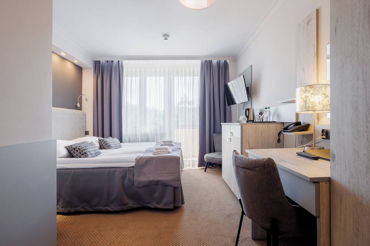 Habitación de hotel con cama, escritorio, silla, televisor y un amplio ventanal con cortinas.